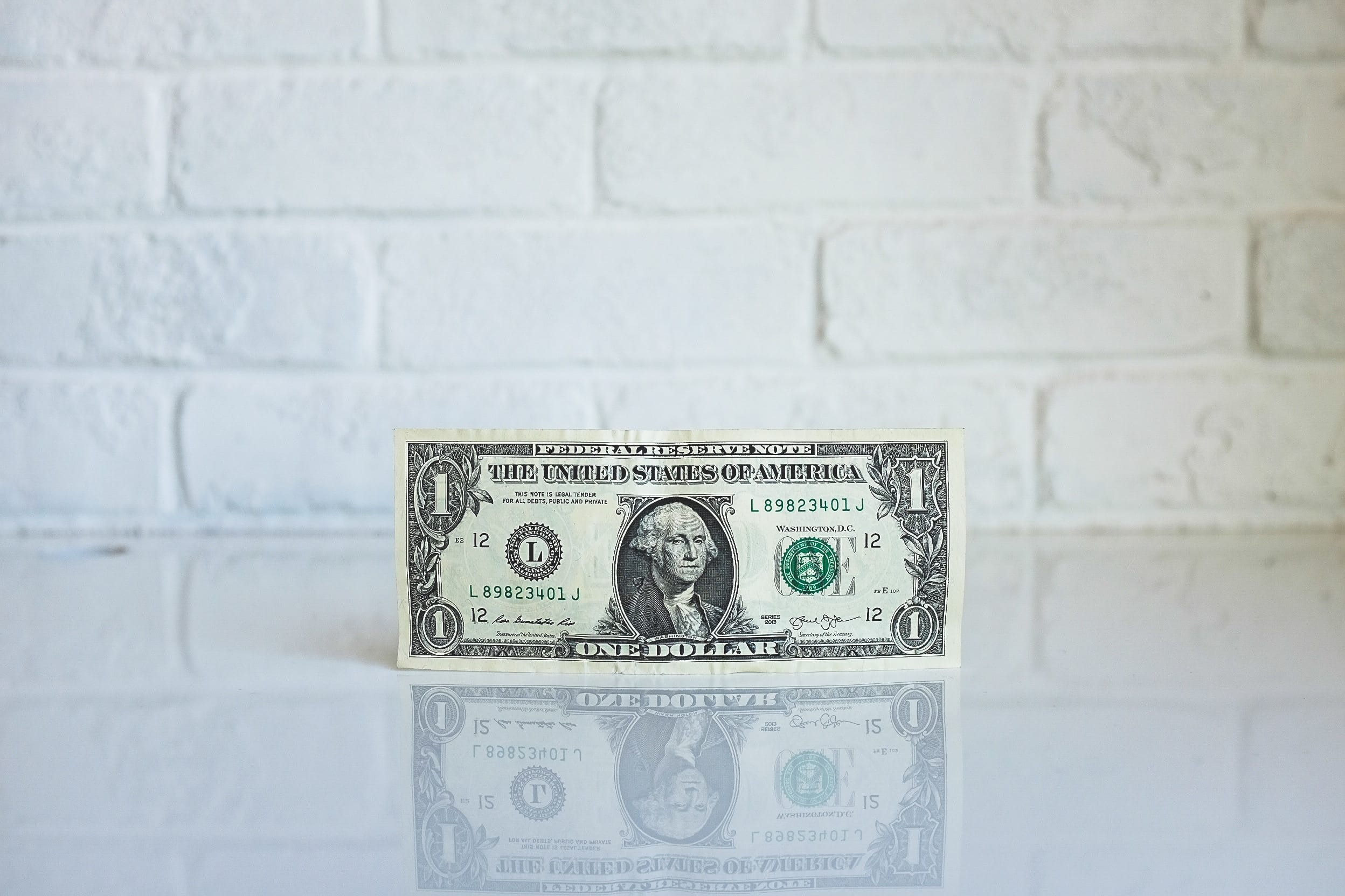 A one dollar bill standing on edge on a reflective white surface with a white brick wall as backdrop; image by NeONBRAND, via Unsplash.com.