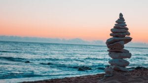 A stack of balanced stones by the water at the beach at sunset; image by Thomas Rey, via Unsplash.com.