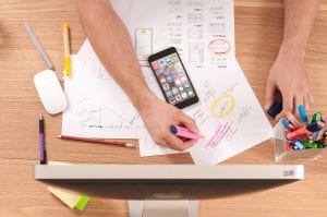 Man in front of computer monitor, with smartphone, writing on white paper; image by <a href="https://unsplash.com/@firmbee">William Iven</a>, via <a href="https://unsplash.com/photos/gcsNOsPEXfs">Unsplash.com</a>.