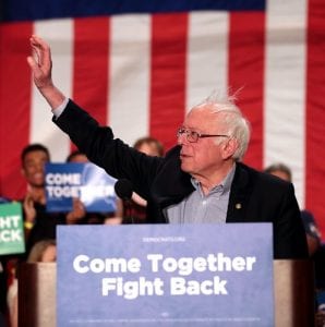 Bernie Sanders, standing at a podium with his right arm upraised. A sign on the podium reads, "Come Together, Fight Back."