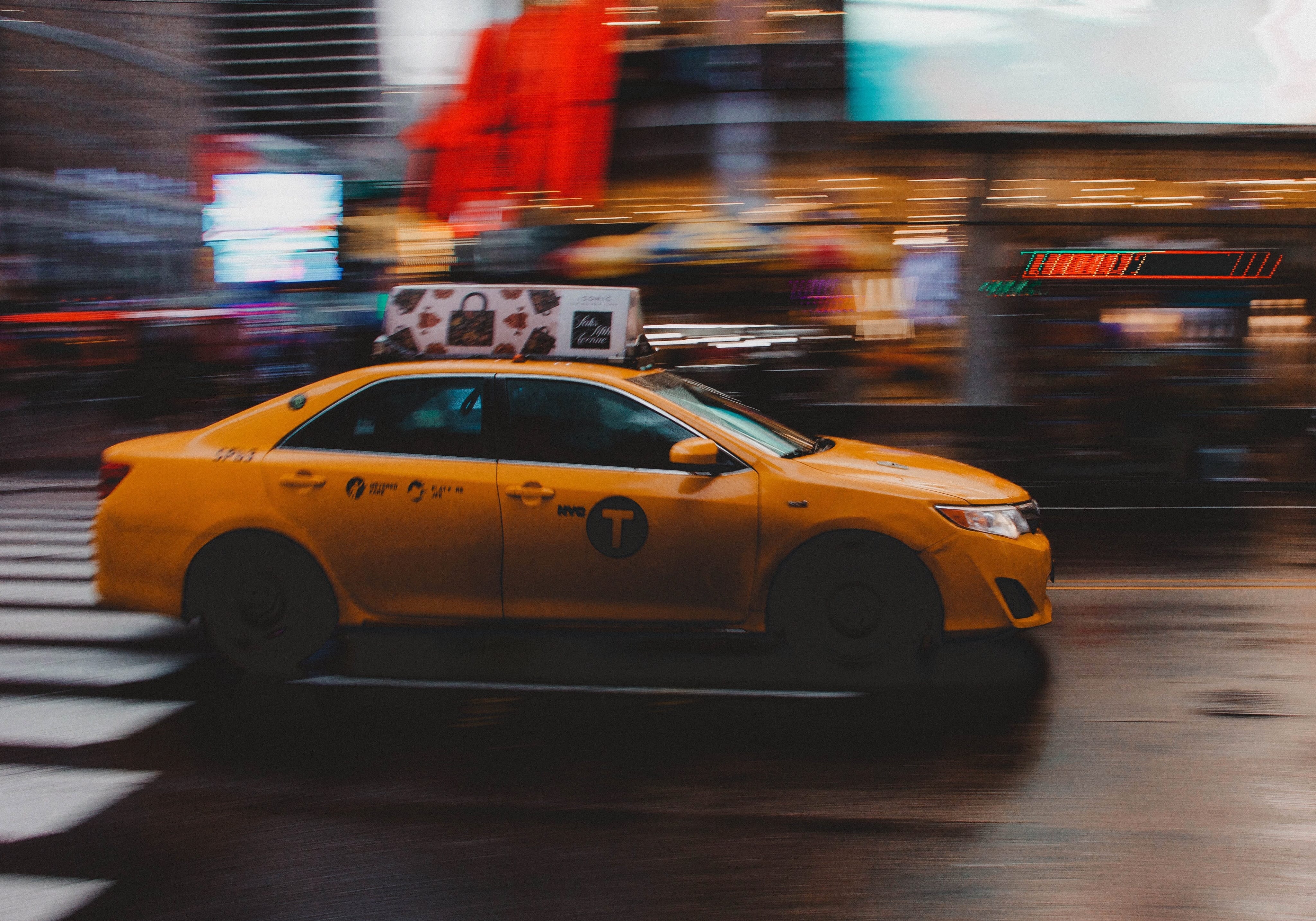 Yellow taxi driving down street, scenery blurred; image by @gehartyler, via Unsplash.com.