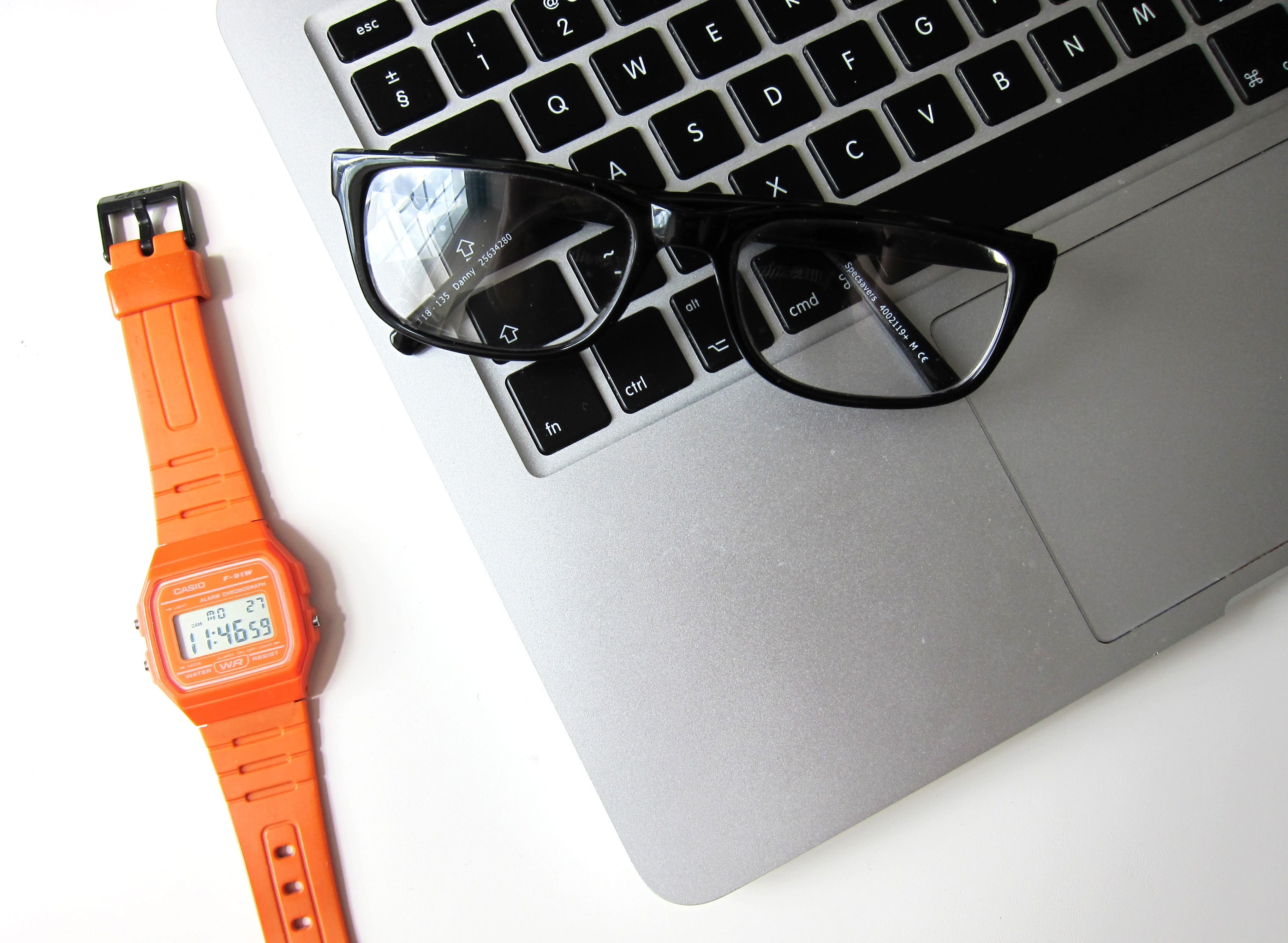 Reading glasses folded on a laptop along with a watch with orange band on a white desk; image by Hello I’m Nik, via Unsplash.com.