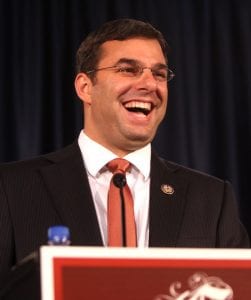 Justin Amash, wearing a dark suit, white shirt, and red tie, stands behind a podium with a wide, laughing smile on his face.