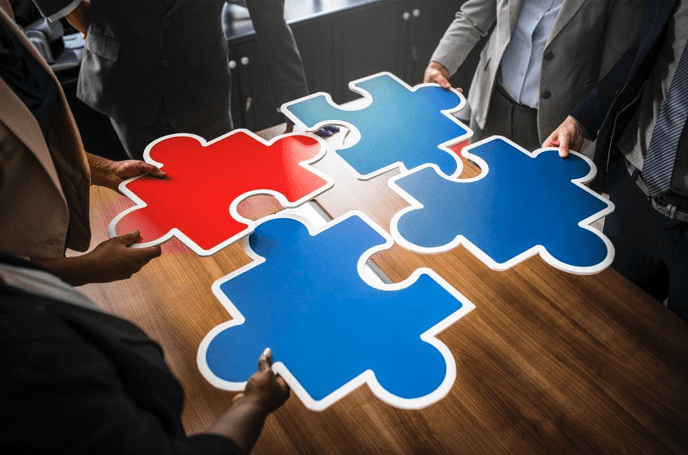 People holding large, multi-colored puzzle pieces over a table, trying to fit them together; image by rawpixels.com, via pexels.com.