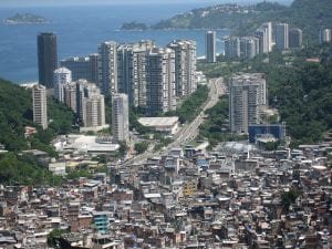 A massive shanty town of makeshift hones and unnamed streets lies against a glittering big city and oceanfront background.