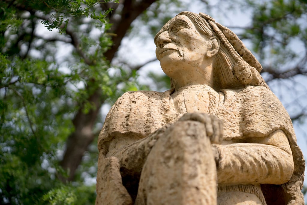 A stone statue of a Native American man looking into the distance, with a tree in the background.
