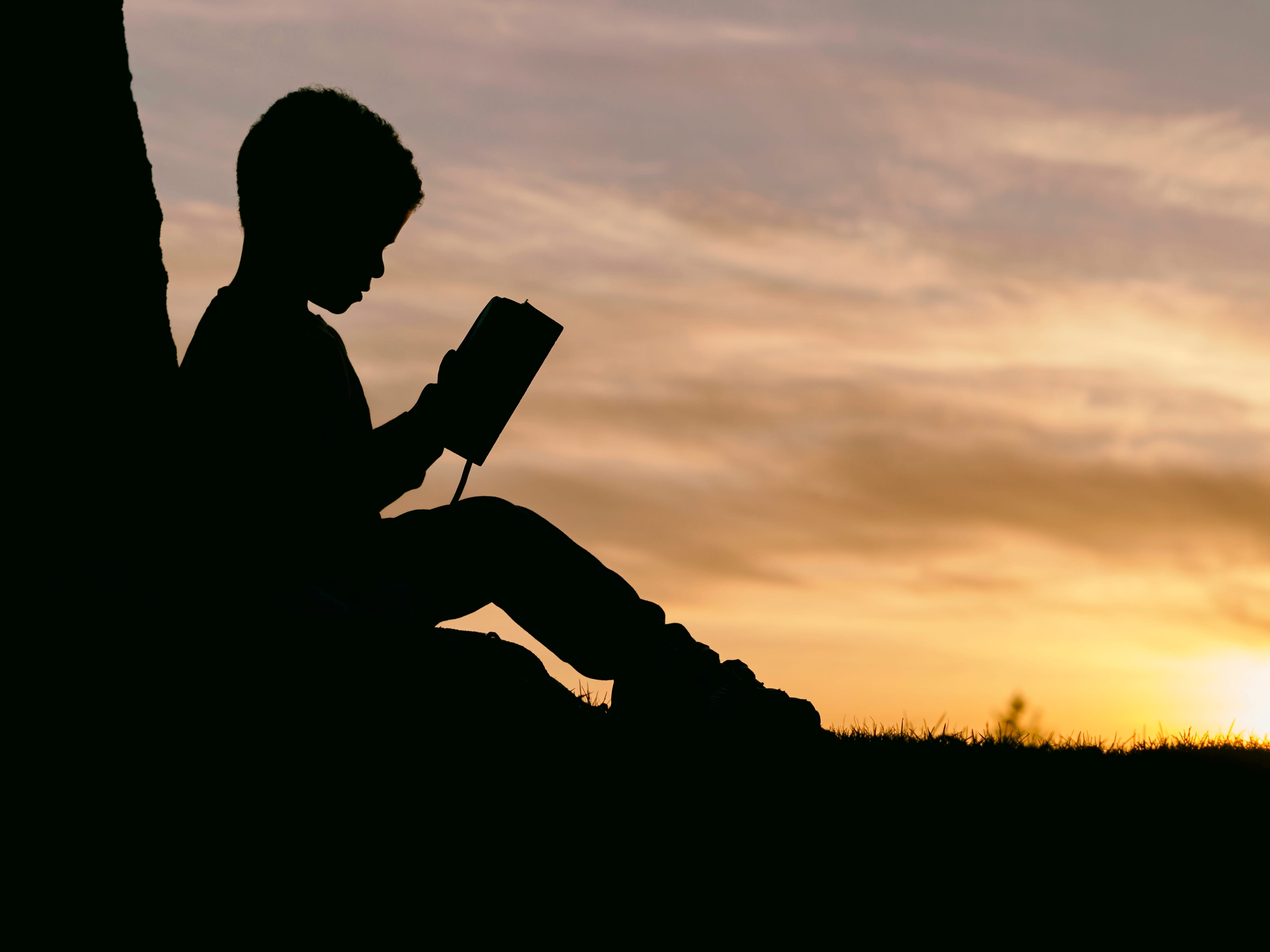 Silhouette of boy leaning against a tree at sunset, reading a book; image by Aaron Burden, via Unsplash.com.