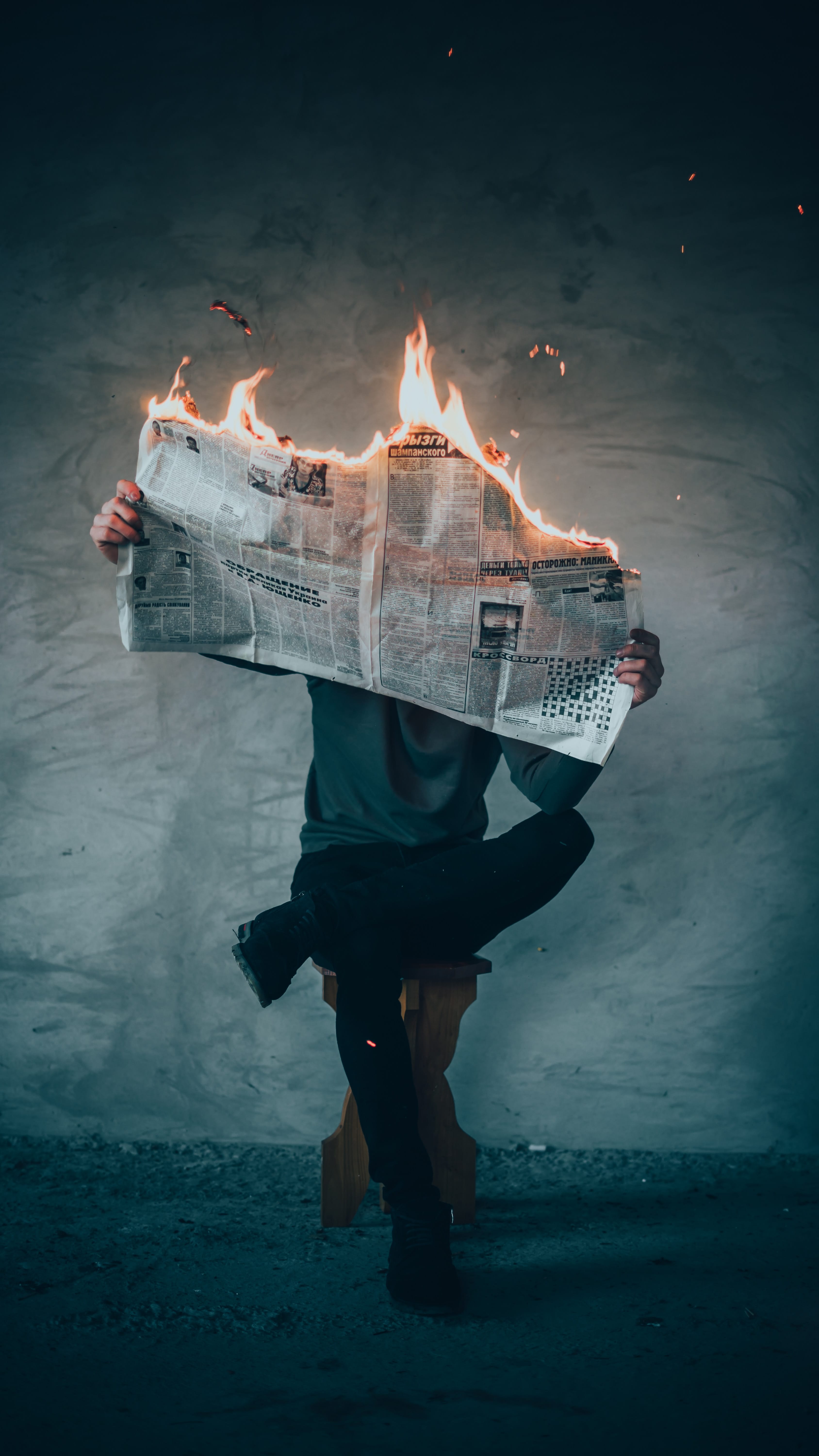 Man sitting on stool reading a newspaper that is on fire; image by Elijah O’Donnell, via Unsplash.com.