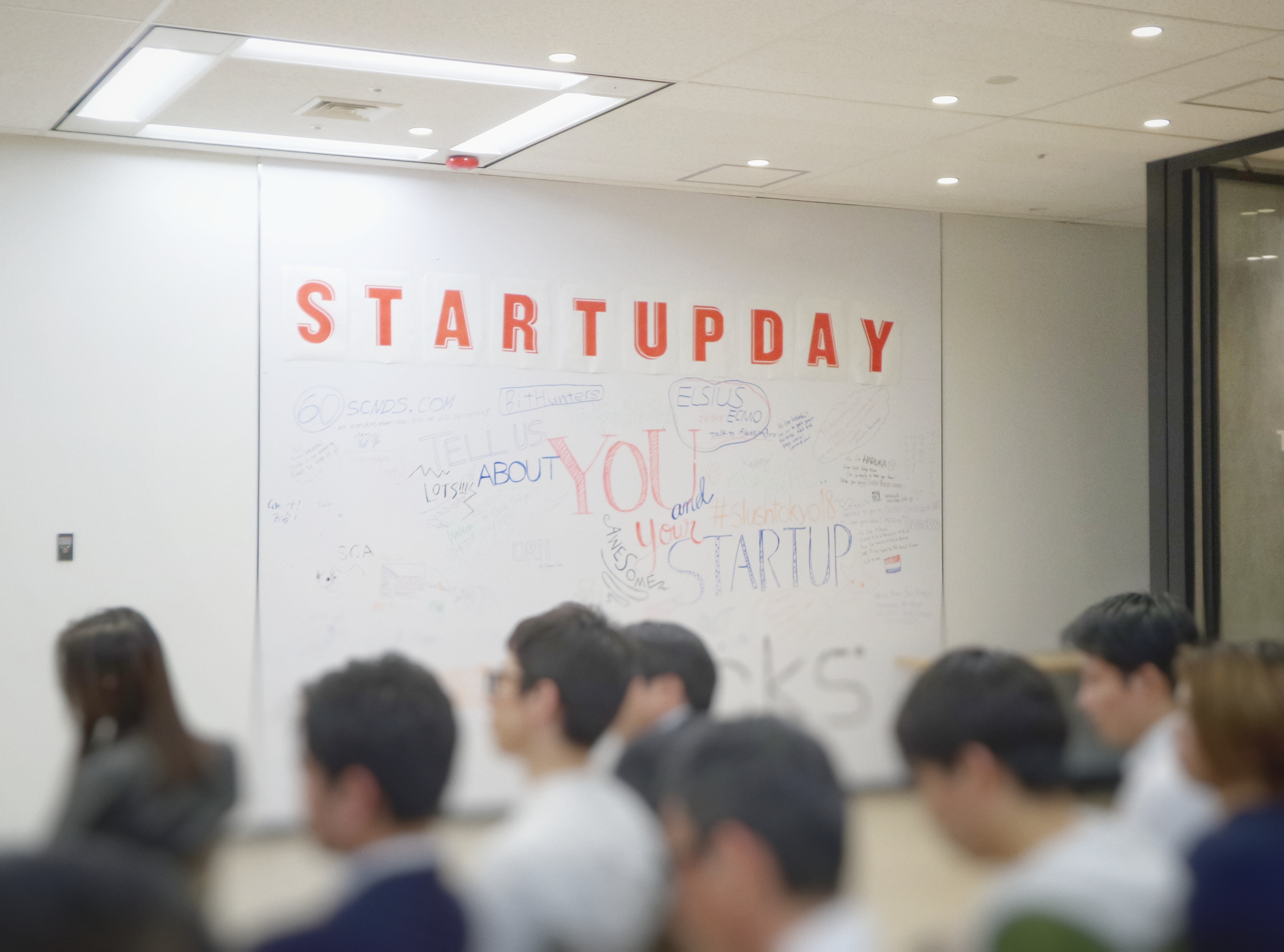 Soft focus of a group of people in a conference room with a white board saying “Startupday.” Image by Franck V., via Unsplash.com.