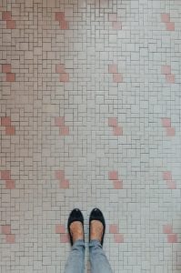 Woman’s feet and legs, in black shoes and jeans, on pink and white tile; image by Kelly Sikkema, via Unsplash.com.