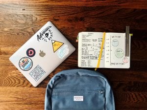 Laptop with stickers on it, blue backpack, and notebook on a brown woodgrain desk; image by Matt Ragland, via Unsplash.com.