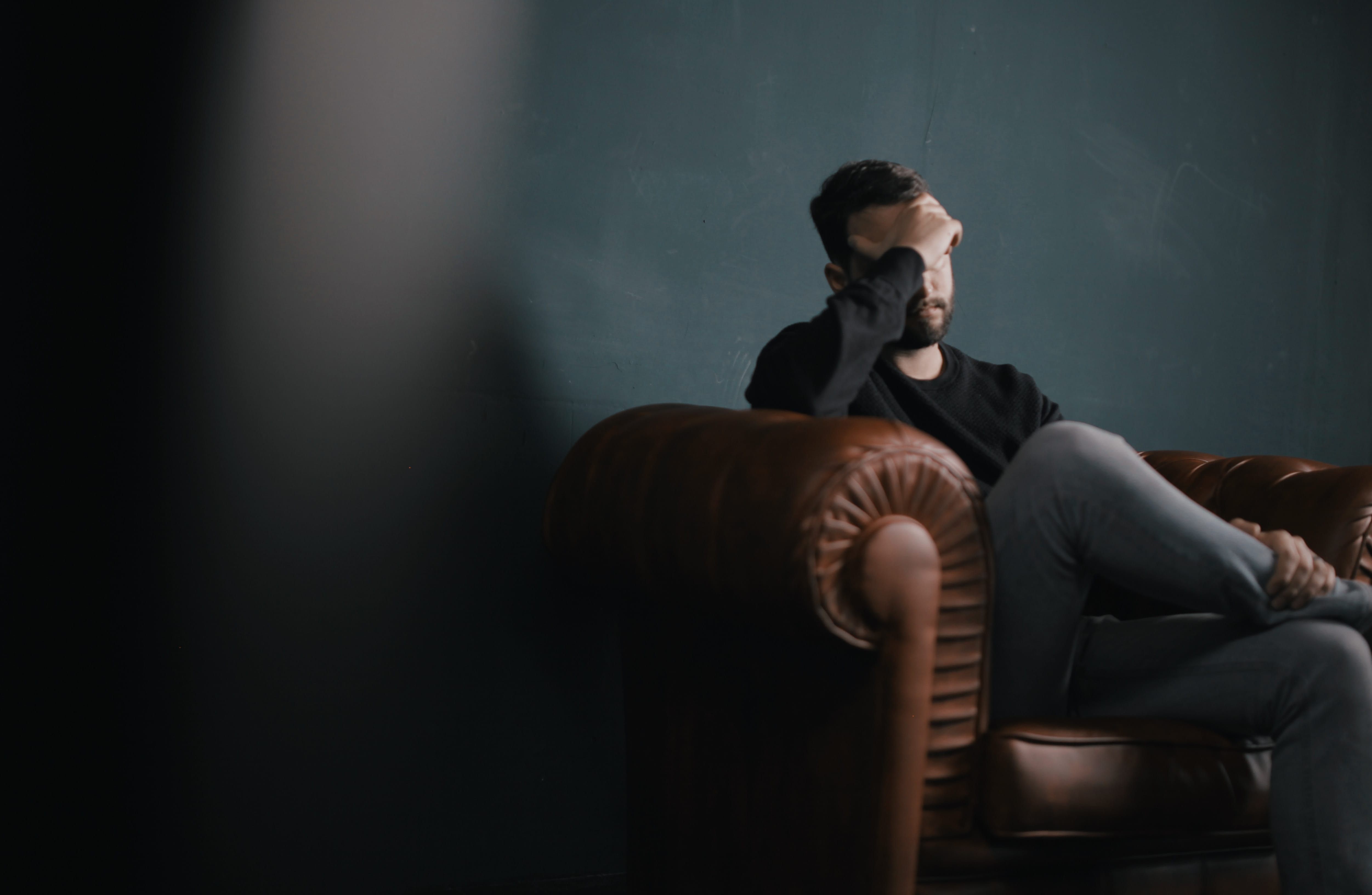 Man sitting on brown leather sofa covering his eyes with one hand; image by Nik Shuliahin, via Unsplash.com.