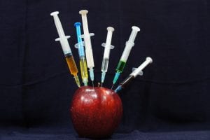 Red apple with multiple fluid-containing syringes stuck into it; image by Sara Bakhshi, via Unsplash.com.