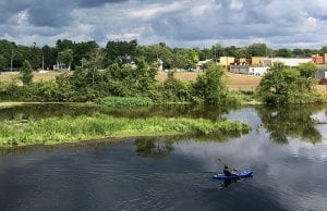 A kayaker paddles through a calm, serene river with plenty of green trees and water plants nearby.
