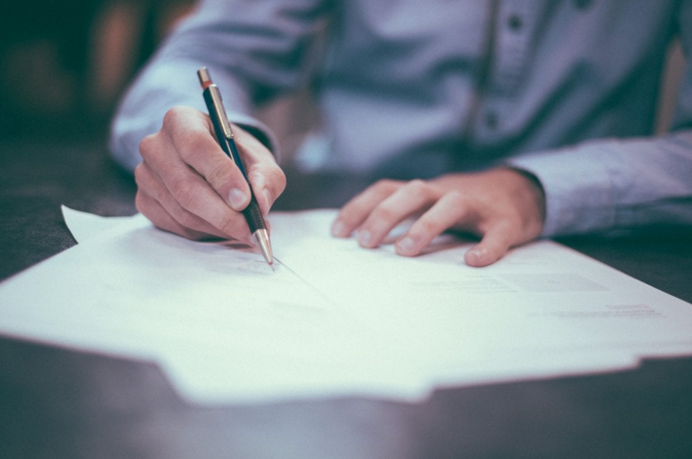 Man in blue dress shirt writing on paperwork; image by Helloquence, via Unsplash.com.