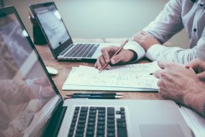 Two men with laptops reviewing paperwork; image by Helloquence, via Unsplash.com.