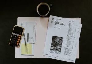 Tax withholding and estimated tax paperwork near a smartphone calculator, pen, paper, and coffee; image by Kelly Sikkema, via Unsplash.com.