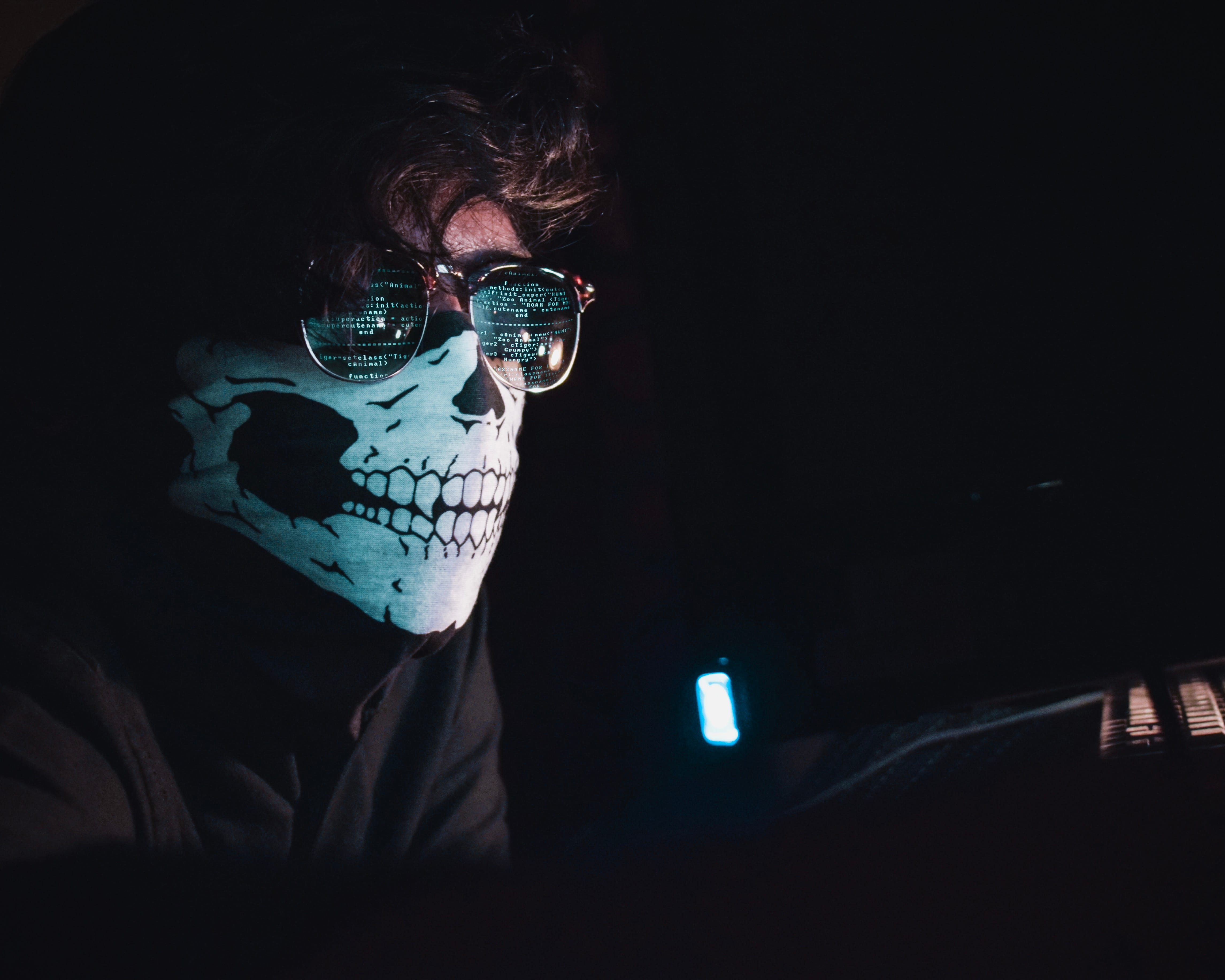 Man wearing a skull mask and reflective glasses in a dark room. Computer code is reflected in his glasses. Image by Nahel Abdul Nadi, via Unsplash.com.