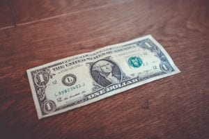 A one-dollar bill on a brown wooden table; image by NeONBRAND, via Unsplash.com.