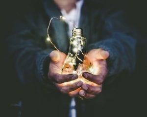 Man holding lit light bulb in outstretched hands; image by Riccardo Annandale, via Unsplash.com.