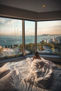 Woman lying on bed facing body of water; image by Roberto Nickson, via Unsplash.com.