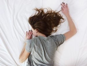 Woman in grey shirt sleeping in bed with white sheets; image by Vladislav Muslakov, via Unsplash.com.
