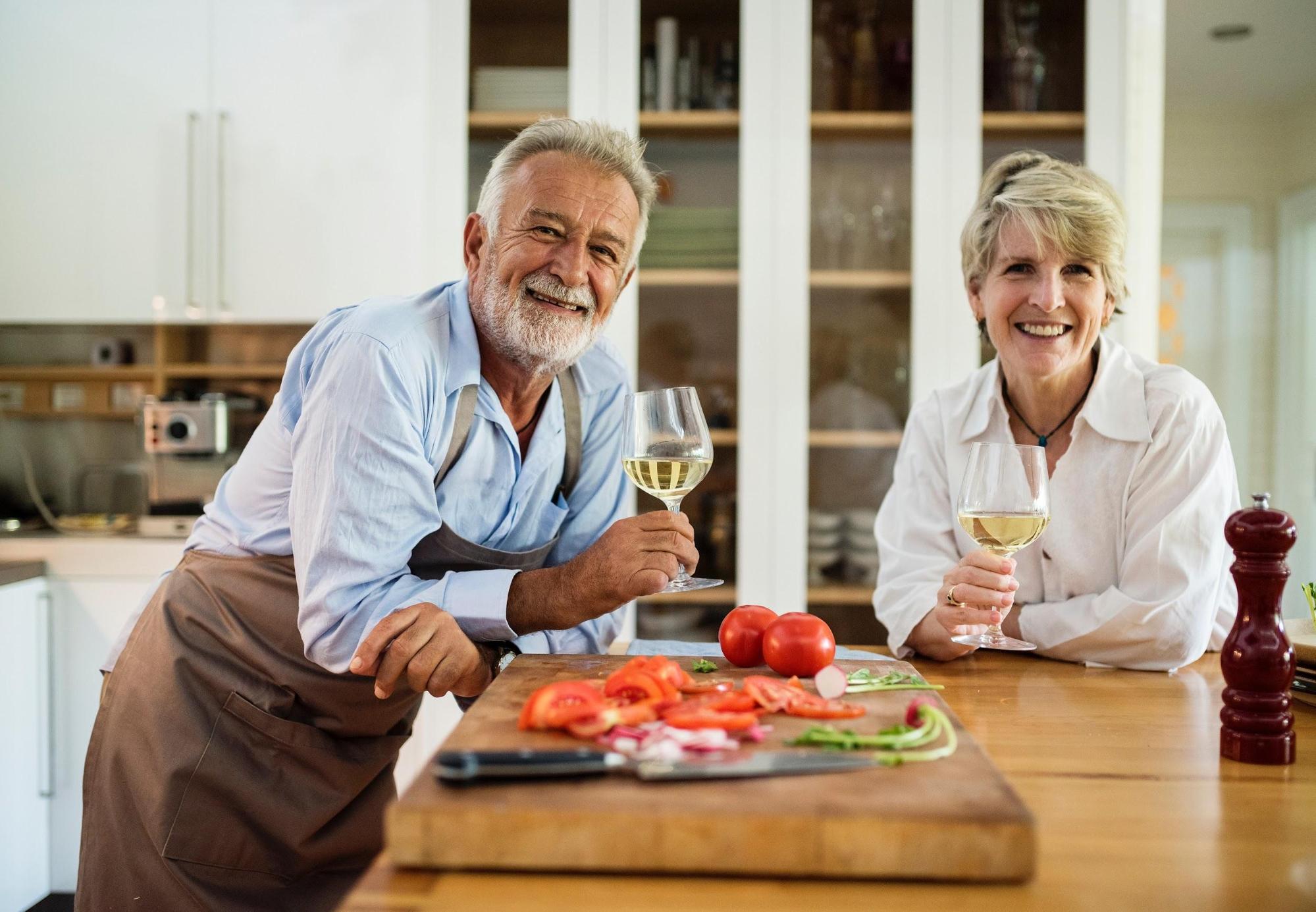 Older man and woman in kitchen with tomatoes on cutting board, drinking wine; image via Pexels.com.