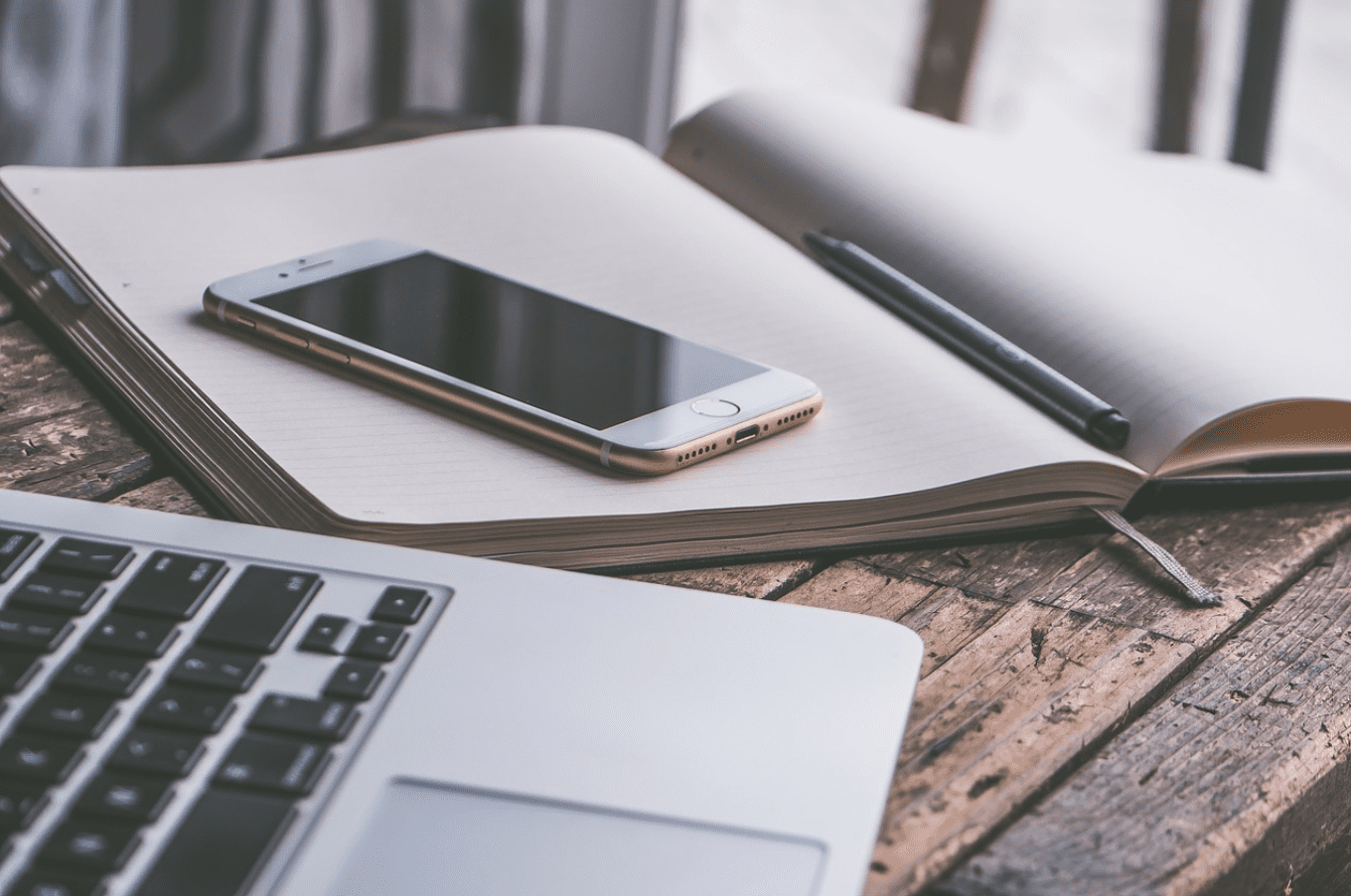 Laptop next to a smartphone sitting on top of an open notebook with a pen, all on a wooden table; image by 6689062, via Pixabay.com.