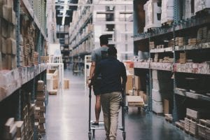 Two men, one on a cart & one pushing it, going through a warehouse; image by Alexander Isreb, via Pexels.com.