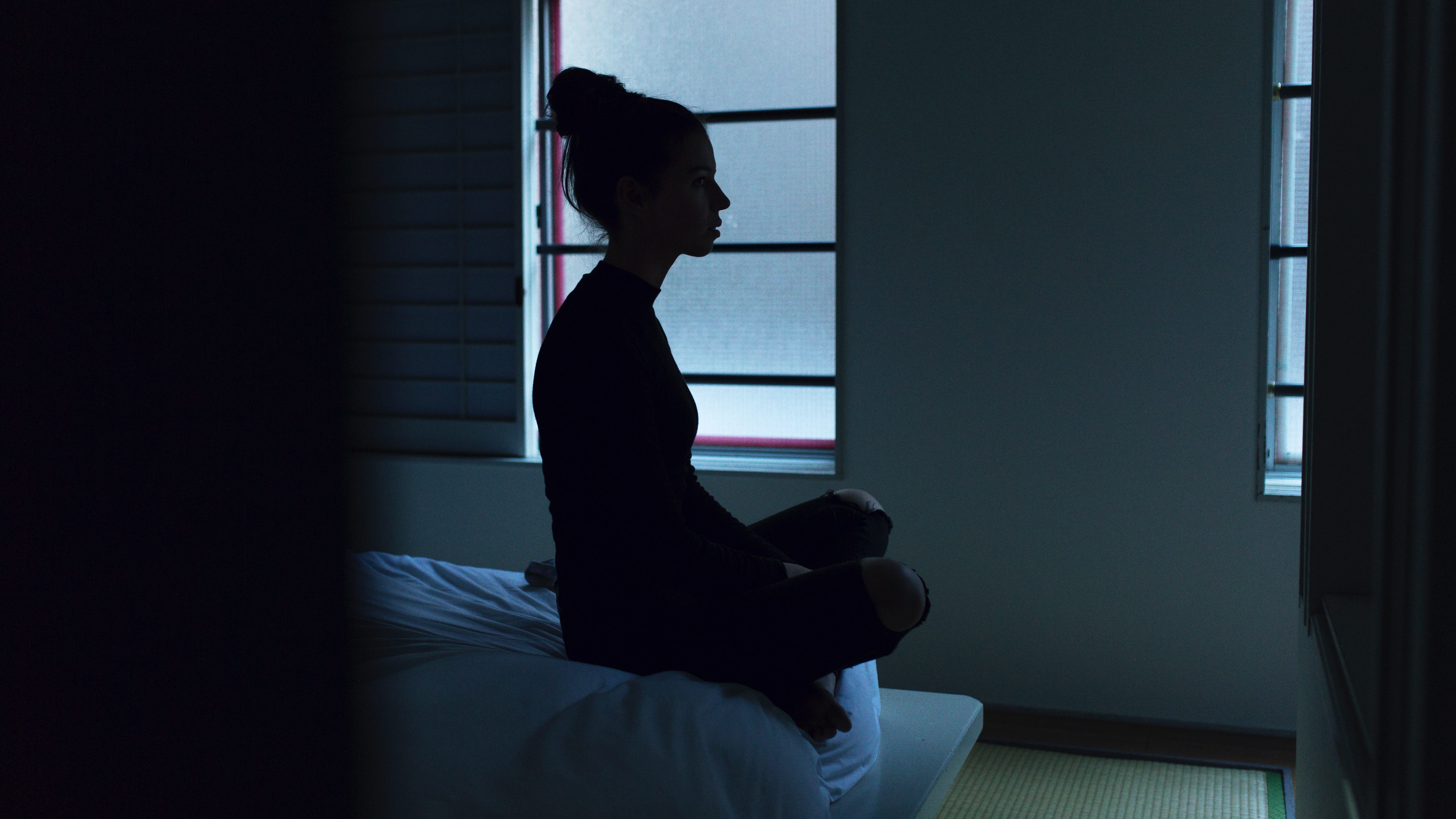 Woman sitting crosslegged on edge of bed in dark room; image by Ben Blennerhassett, via Unsplash.com.