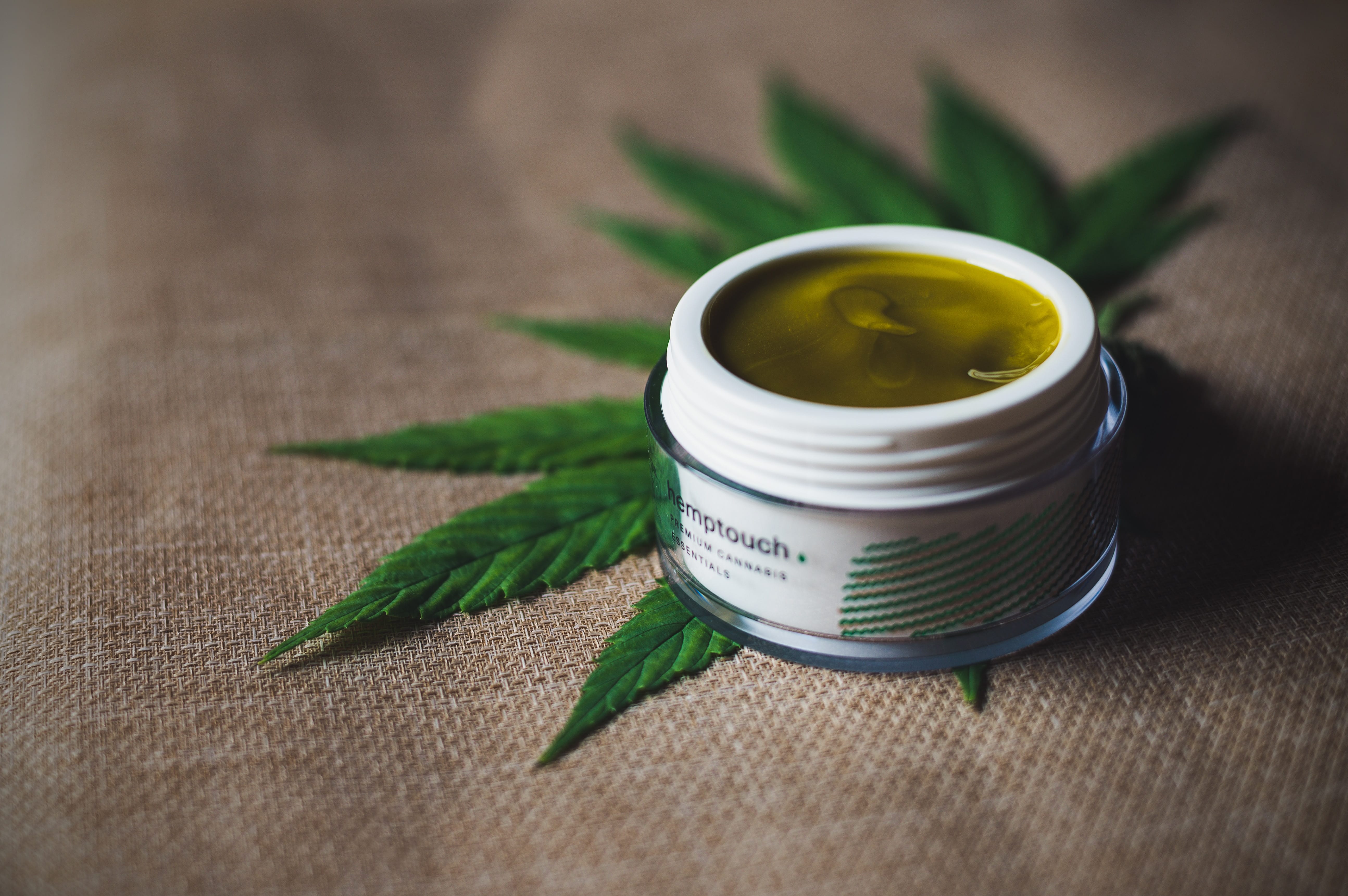 White container of CBD on top of hemp leaf, both on a burlap surface; image by CBD Infos, via Unsplash.com.