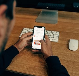 Man sitting at desk with keyboard, mouse, and monitor, while using Twitter on a smartphone; image by Charles, via Unsplash.com.