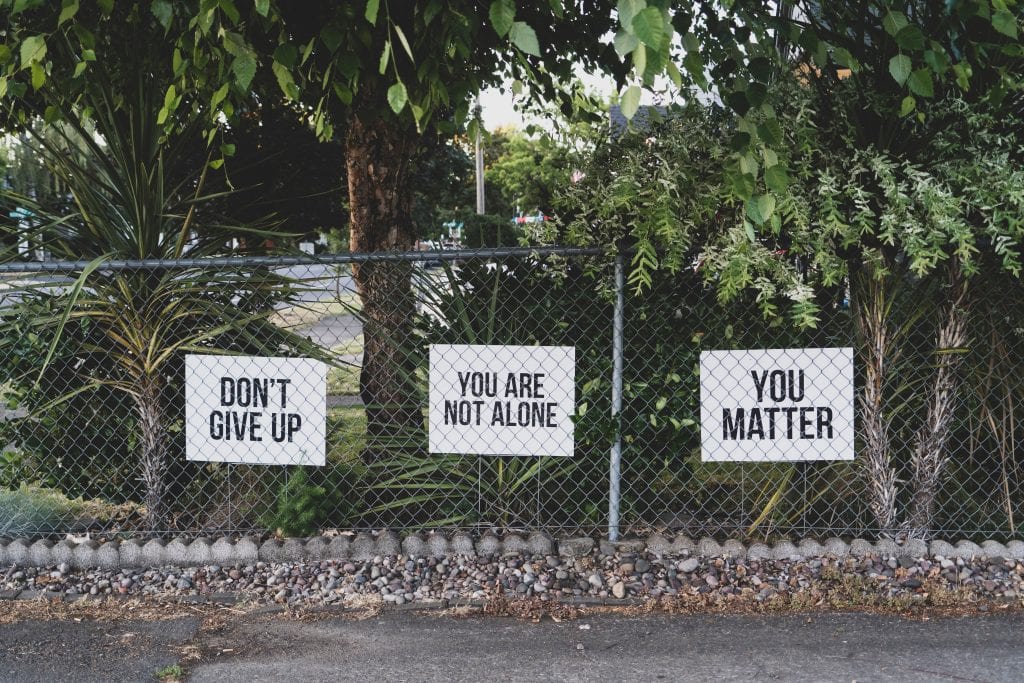 Three signs on a chain link fence, reading (in order) “Don’t Give Up,” “You Are Not Alone,” and “You Matter.” Image by Dan Meyers, via Unsplash.com.