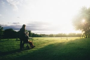 Woman standing next to woman riding wheelchair on grass in the sun; image by Dominik Lange, via Unsplash.com.