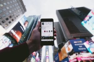 Man holding a smartphone toward the sky with tall buildings in the background; image by Jakob Owens, via Unsplash.com.