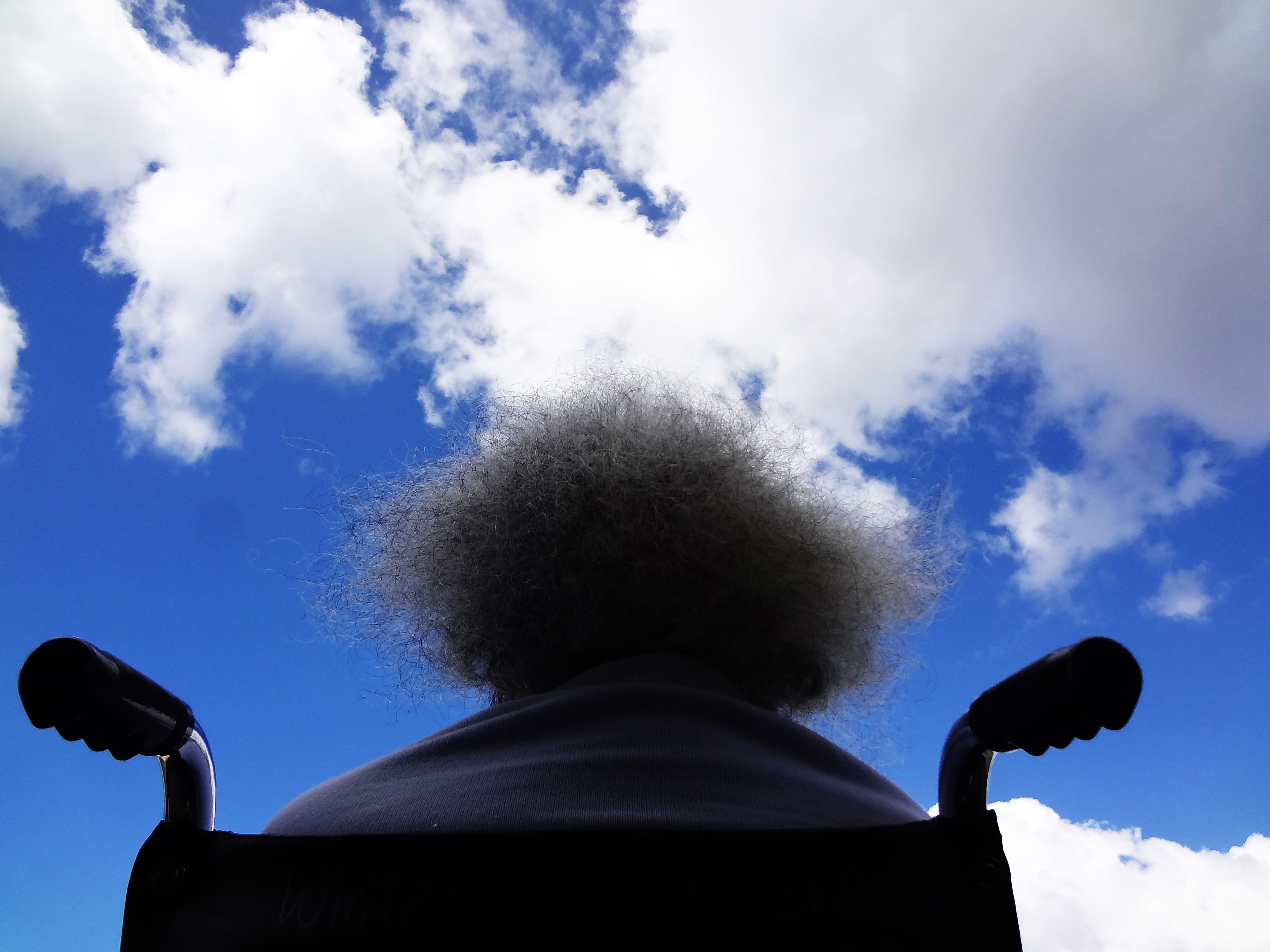 Rear view of woman in wheelchair looking at blue sky with clouds; image by James Williams, via Unsplash.com.