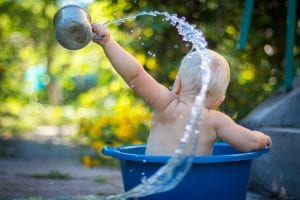 Baby in blue plastic tub flinging water with cup; image by Lubomirkin, via Unsplash.com.