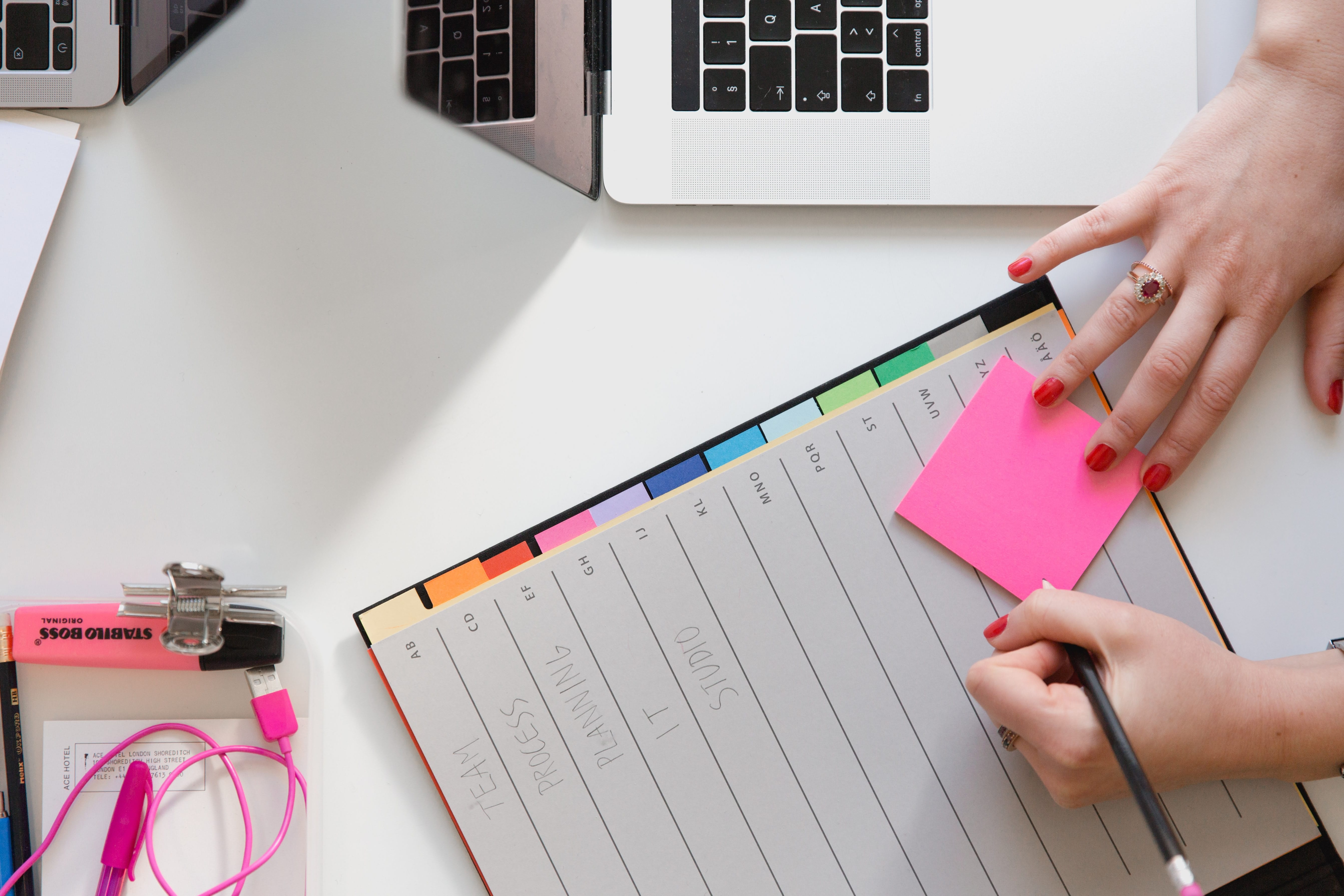Woman holding pencil and pink sticky note next to laptop and planner; image by Marten Bjork, via Unsplash.com.