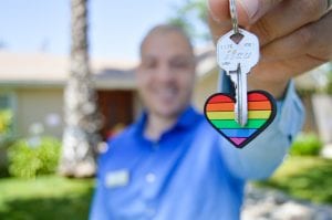 Man in blue shirt holding out key on heart shaped, rainbow keyring; image by Maurice Williams, via Unsplash.com.
