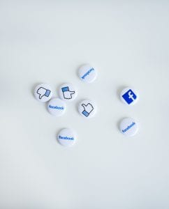 White button pins with blue printing showing “Facebook,” the Facebook logo, and the thumbs up for likes; image by NeONBRAND, via Unsplash.com.