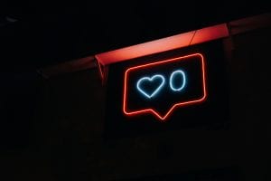 Red and blue neon sign showing a comment bubble and zero likes against a black background; image by Prateek Katyal, via Unsplash.com.