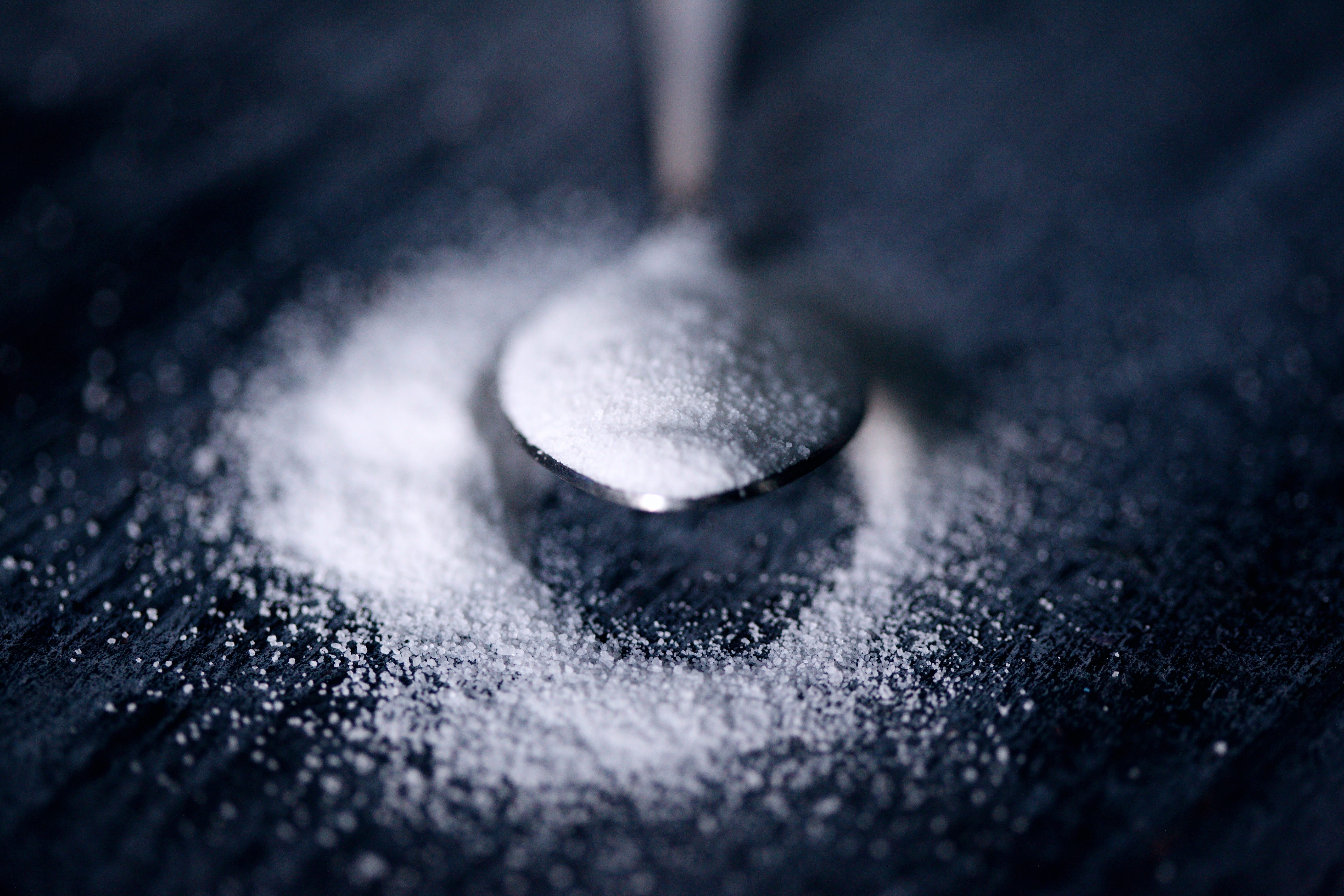 Teaspoon full of sugar on black surface with some spread around; image by Sharon McCutcheon, via Unsplash.com.