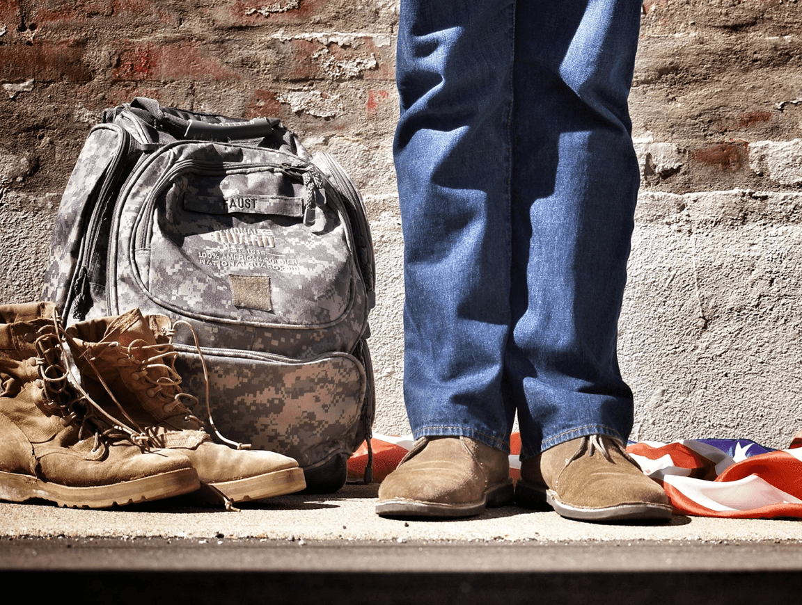 Veteran in blue jeans standing by military backpack, boots, and an American Flag; image by Benjamin Faust, via Unsplash.com.