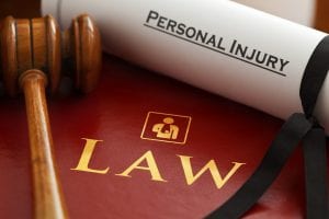 Gavel and rolled paper with “Personal Injury” on it sitting on top of dark red binder titled “Law.” Image by Claimaccident, via pixabay.com. No changes.