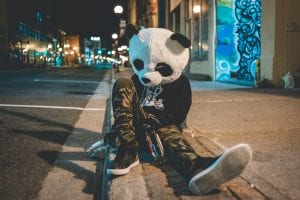 Man in camp pants, dark sweatshirt, and panda costume head slumped against street sign with alcohol bottles in both hands; image by Eric Mclean, via Unsplash.com.