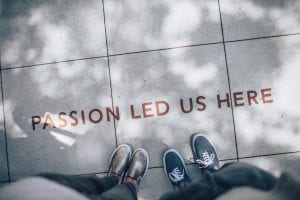 Two people standing on grey tile paving with “Passion Led Us Here” printed at their feet; image by Ian Schneider, via Unsplash.com.