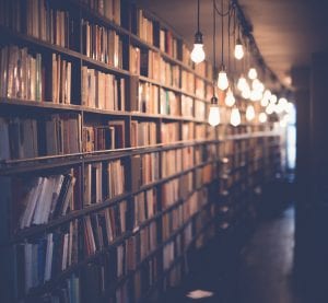 Library shelves lit by hanging bare bulbs; image by Janko Ferlič, via Unsplash.com.