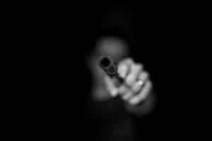Greyscale image of person pointing a gun at viewer; image by Max Kleinen, via Unsplash.com.