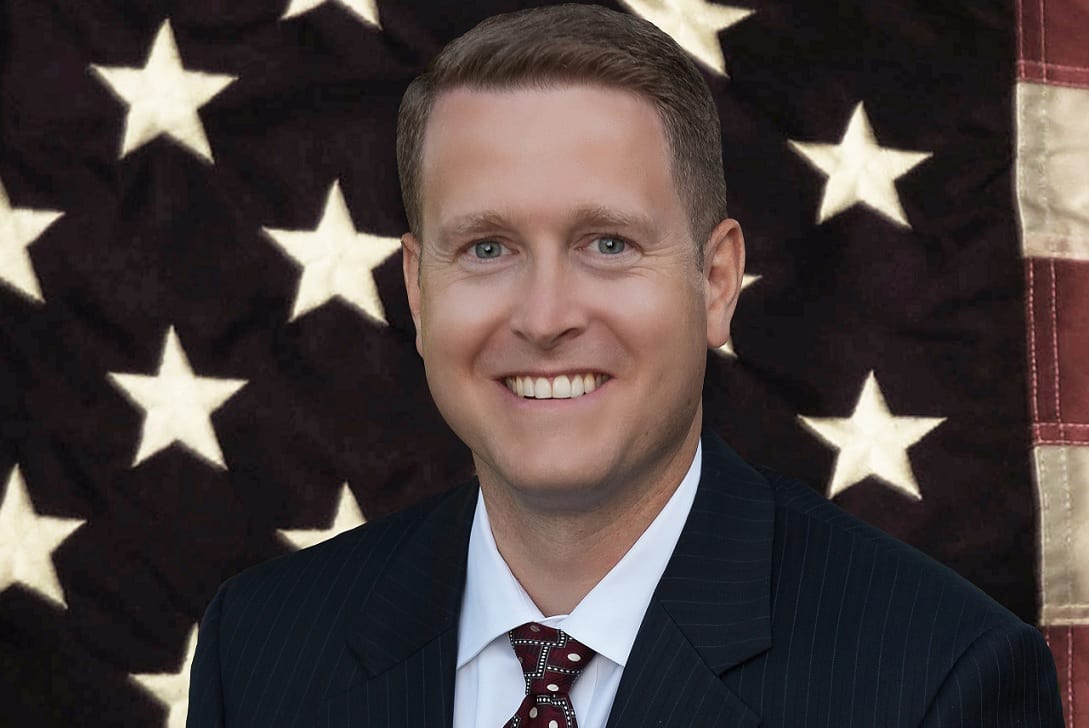 A Caucasian man with short brown hair, wearing a dark suit, standing in front of a large American flag.