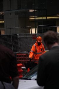 Man wearing reflective suit and hard hat standing beside link fence; image by Dean Bennett, via Unsplash.com.